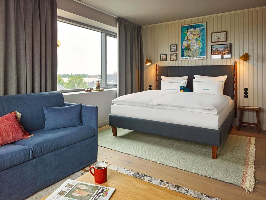 A bedroom with a bed, a blue sofa, a window with curtains, and a view of the outside.