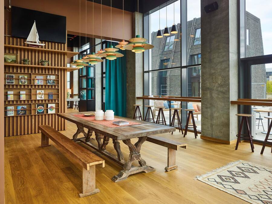 A modern interior with a wooden table, benches, hanging pendant lights, and a bookshelf with books and decorative items.