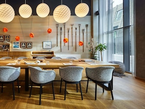 Un espace de restauration moderne avec une longue table en bois, des chaises à revêtement gris, des luminaires sphériques suspendus et un présentoir mural pour les couverts.