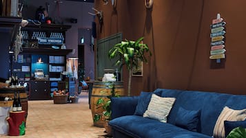 Une pièce chaleureuse avec un canapé bleu, une table basse en bois et une grande plante en pot. Un panneau avec les numéros des chambres et un panneau indiquant le bar sont fixés au mur.