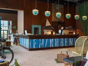 A reception area with a wooden counter, hanging pendant lights, and a view of the outside. - 25hours Hotel Paper Island (Copenhagen)