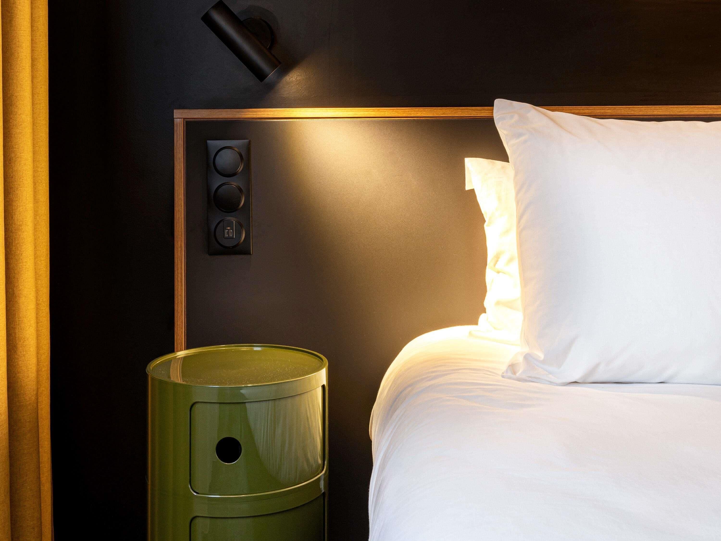 A hotel room with a bed, a nightstand, a lamp, and a control panel.