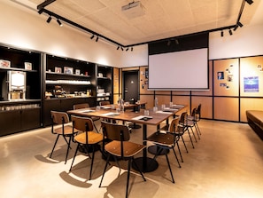 A conference room with a large table, chairs, a projector screen, and a coffee machine. - Ibis Styles Paris Villejuif (Villejuif)