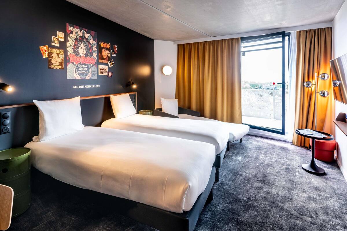 A hotel room with two beds, a large window with curtains, a wall decorated with various posters, and a small table with a lamp.