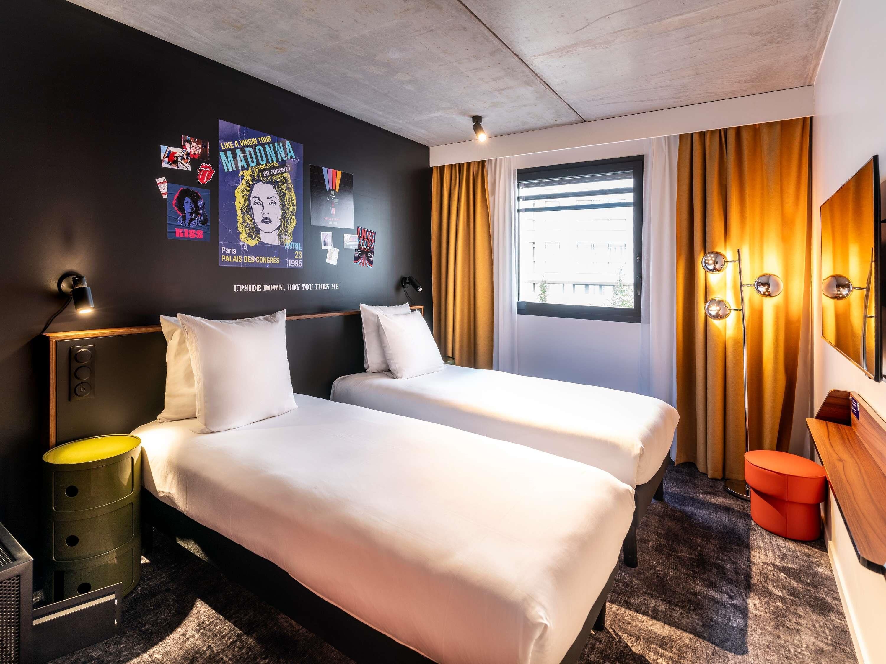 Hotelkamer met twee bedden, een raam met gordijnen, een muur met posters, een bedlamp en een spiegel.