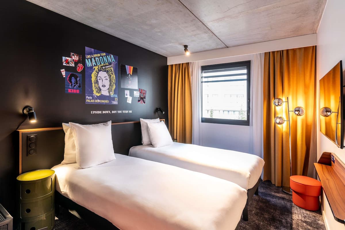 Habitación de hotel con dos camas, una ventana con cortinas, una pared decorada con carteles, una lámpara de noche y un espejo.