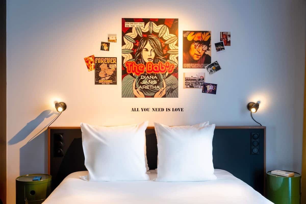 Una cama bien hecha con sábanas blancas, una pared con varios carteles y fotografías, y dos lámparas de noche.