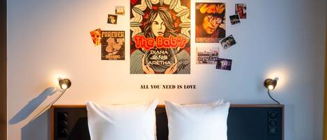 Una cama bien hecha con sábanas blancas, una pared con varios carteles y fotografías, y dos lámparas de noche.