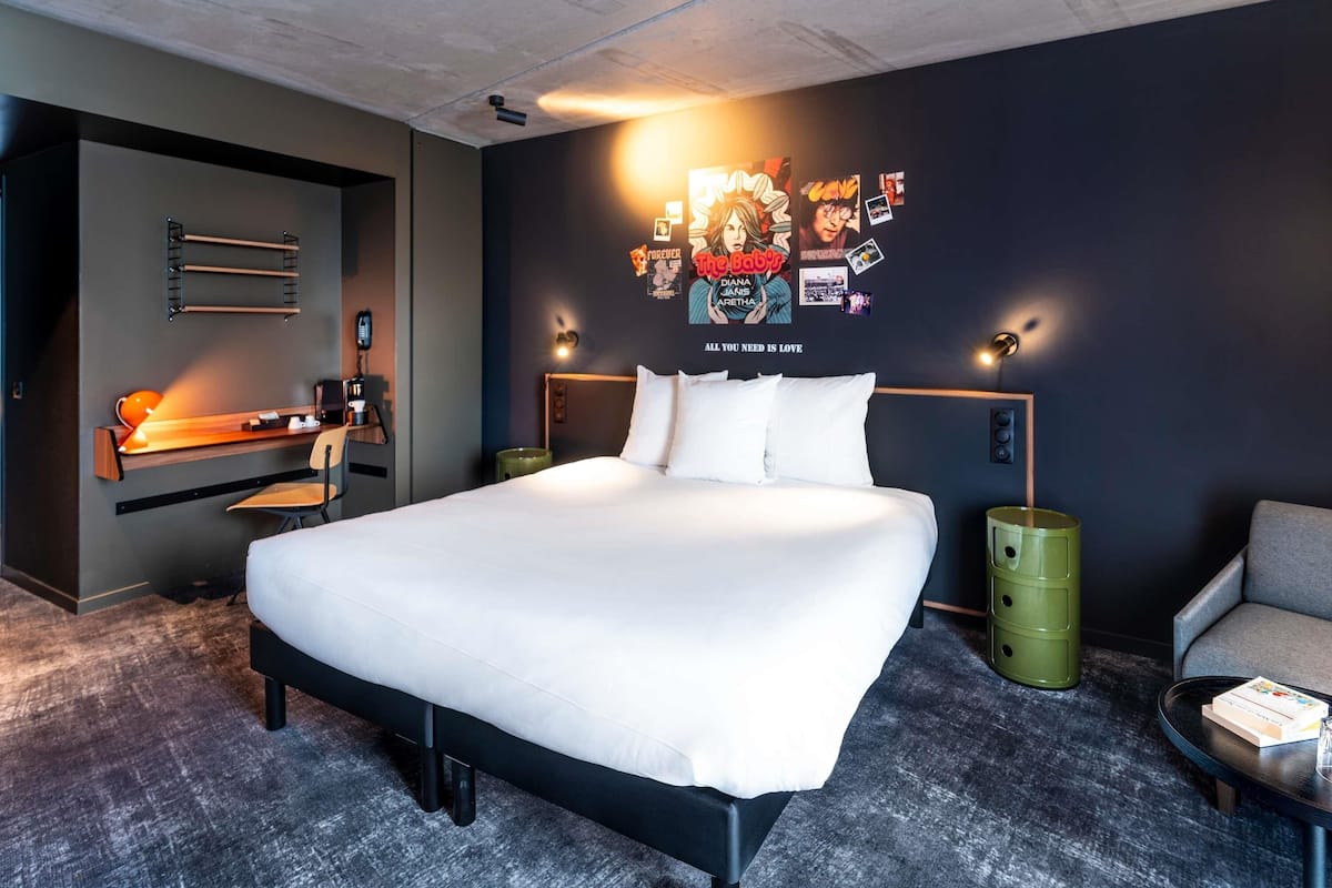 Habitación de hotel moderna con una cama grande, un escritorio con una silla, una mesita con un libro, y una pared decorada con carteles y una cita.