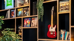 Una estantería con varios libros, una guitarra eléctrica roja y una planta verde.