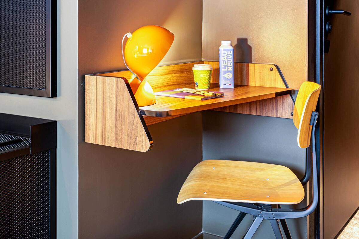 A corner desk with a wooden chair, a yellow lamp, a cup, and a bottle of Le Neuf.