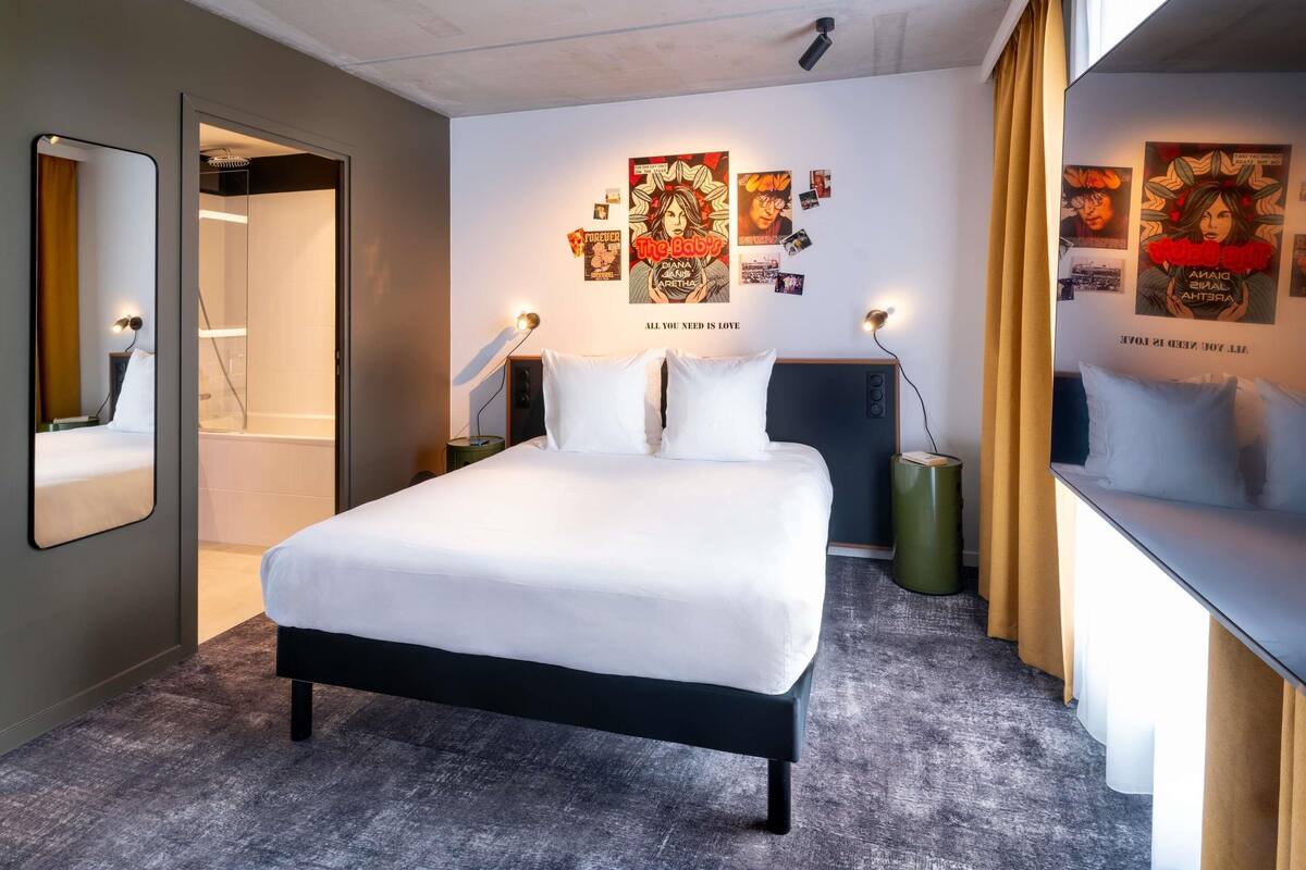 A modern hotel room with a large bed, a mirror, a wall with posters, and a window with curtains.