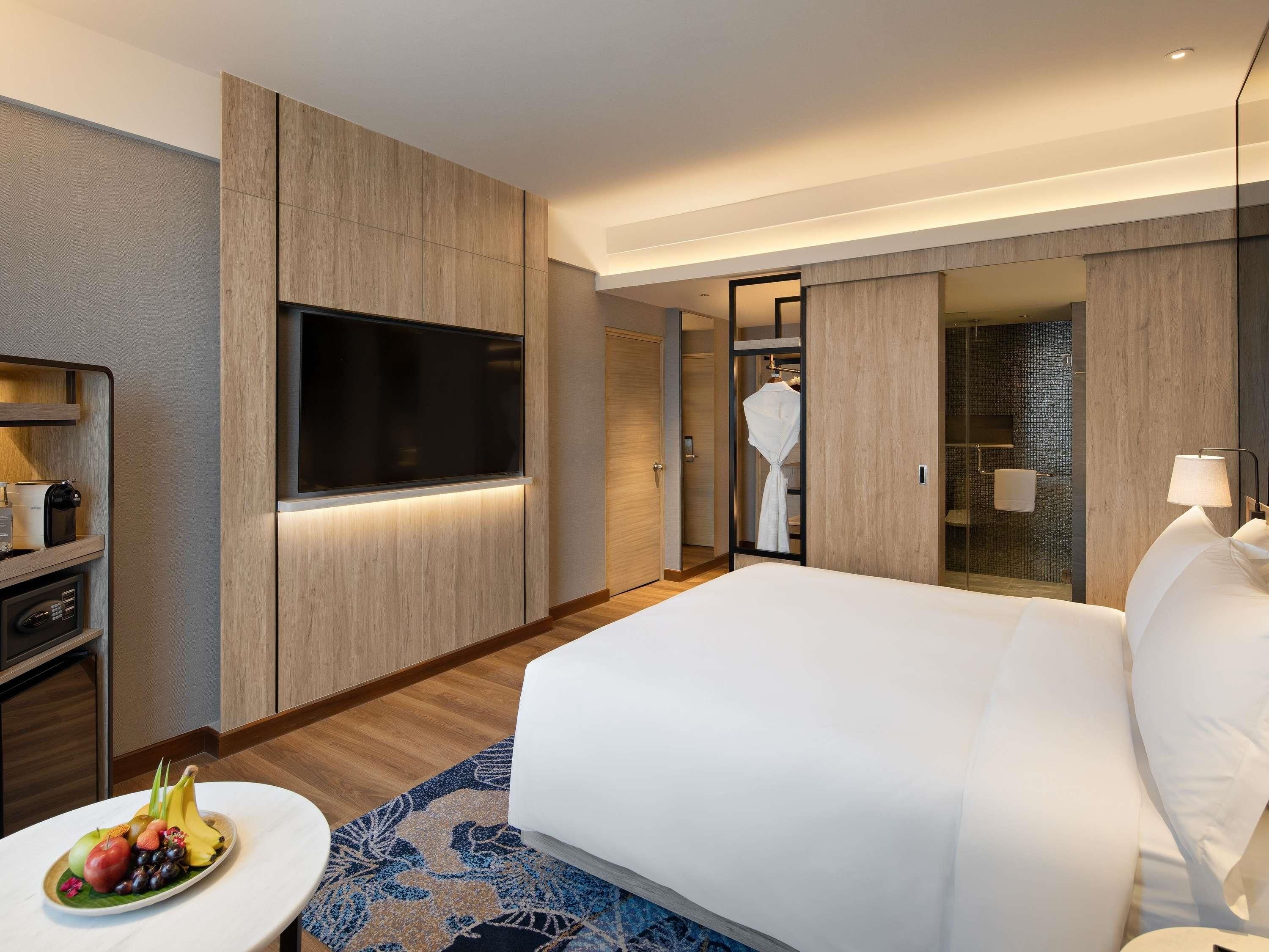A hotel room with a bed, a TV, a desk with a coffee maker, a fruit bowl, and a bathroom visible through an open door.