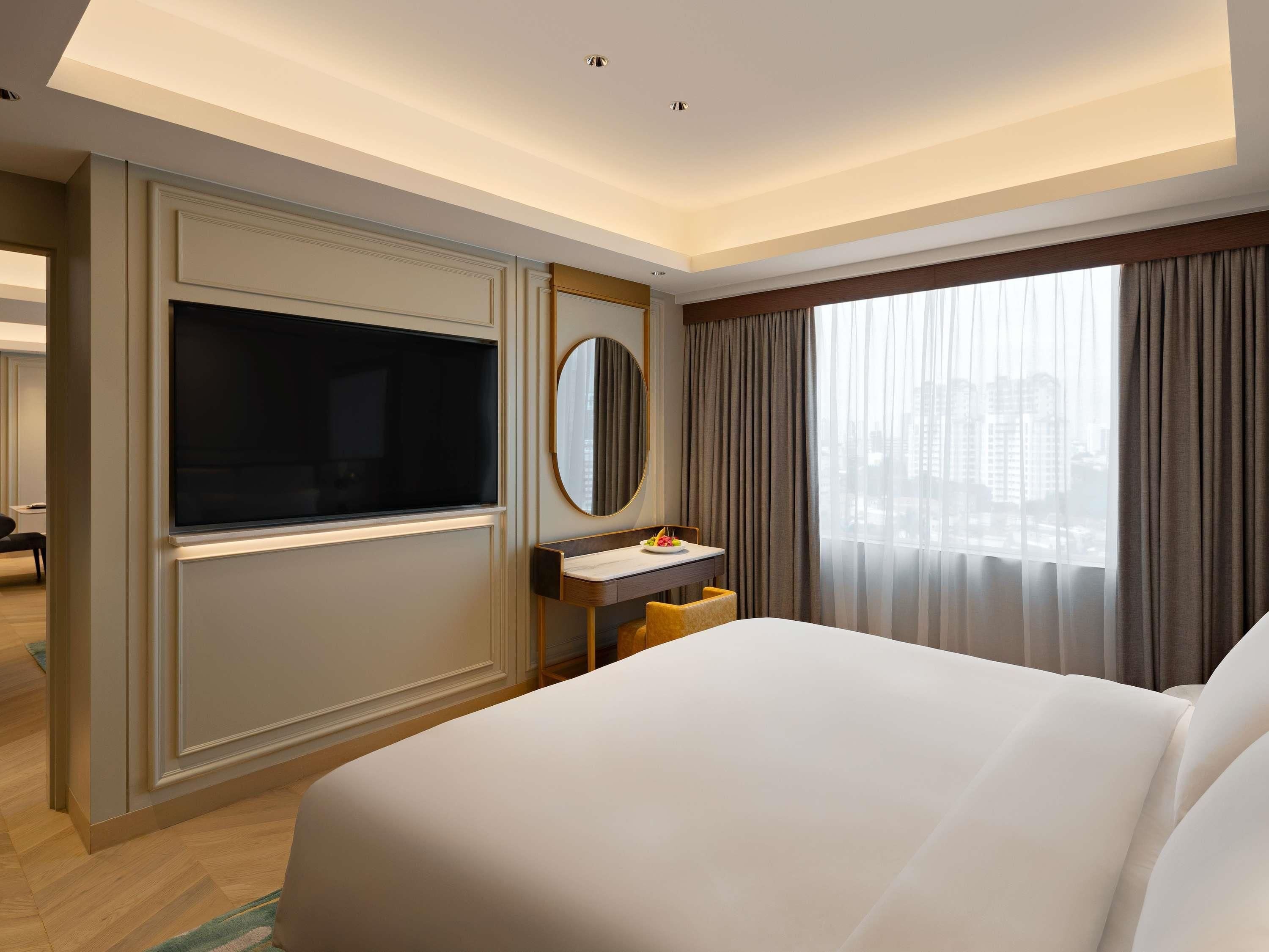 A hotel room with a large bed, a TV mounted on the wall, a small desk with a mirror, and a window with curtains.