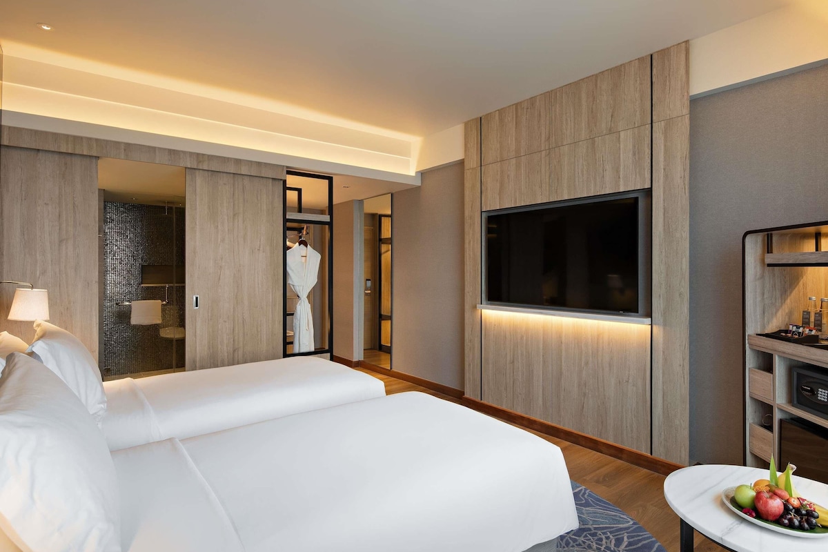 A modern hotel room with a large bed, a TV, a minibar, and a bathroom with a mirror and towel rack.