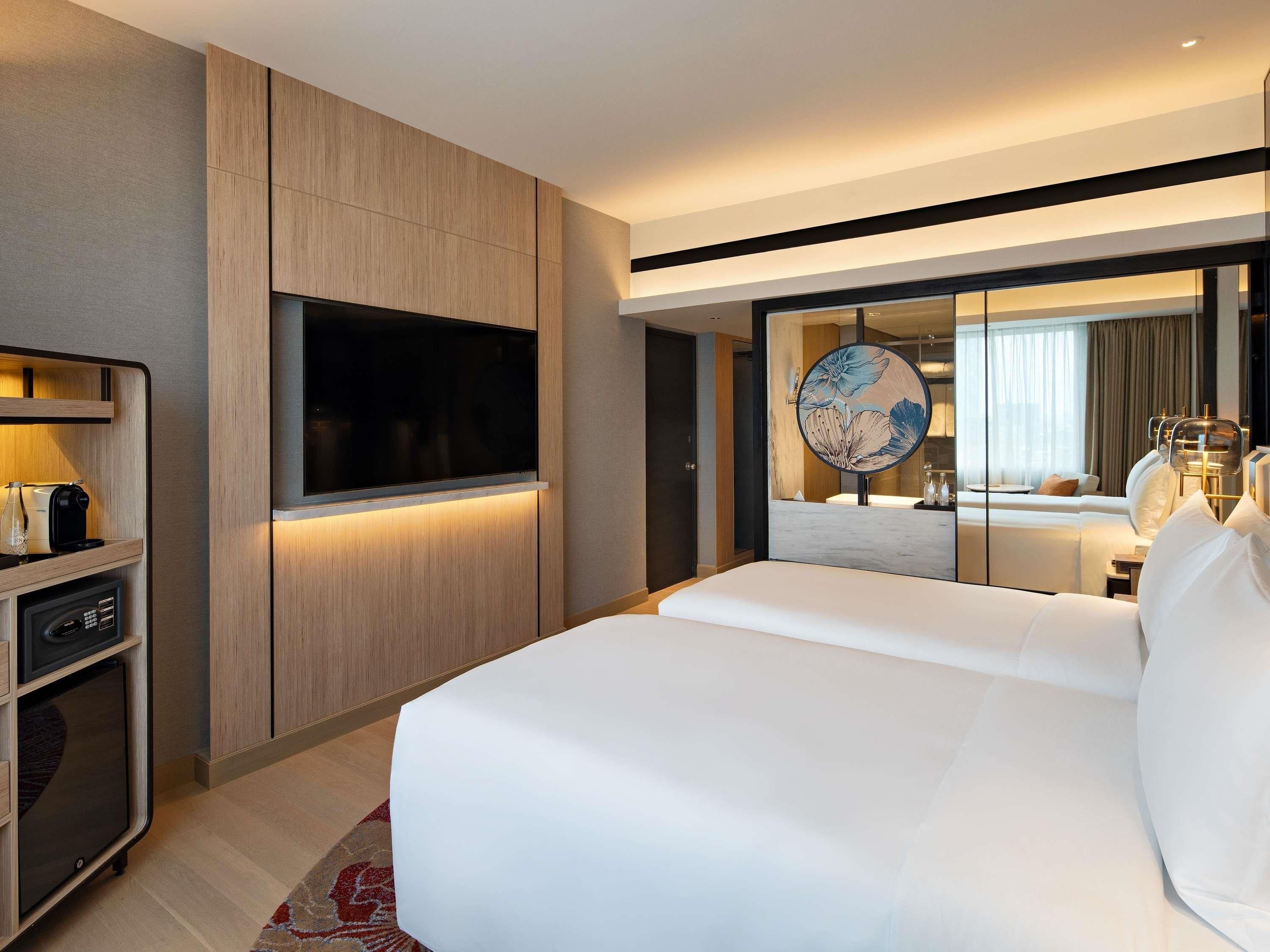 A modern hotel room with a large bed, a flat-screen TV mounted on the wall, a minibar, and a large mirror reflecting the room.