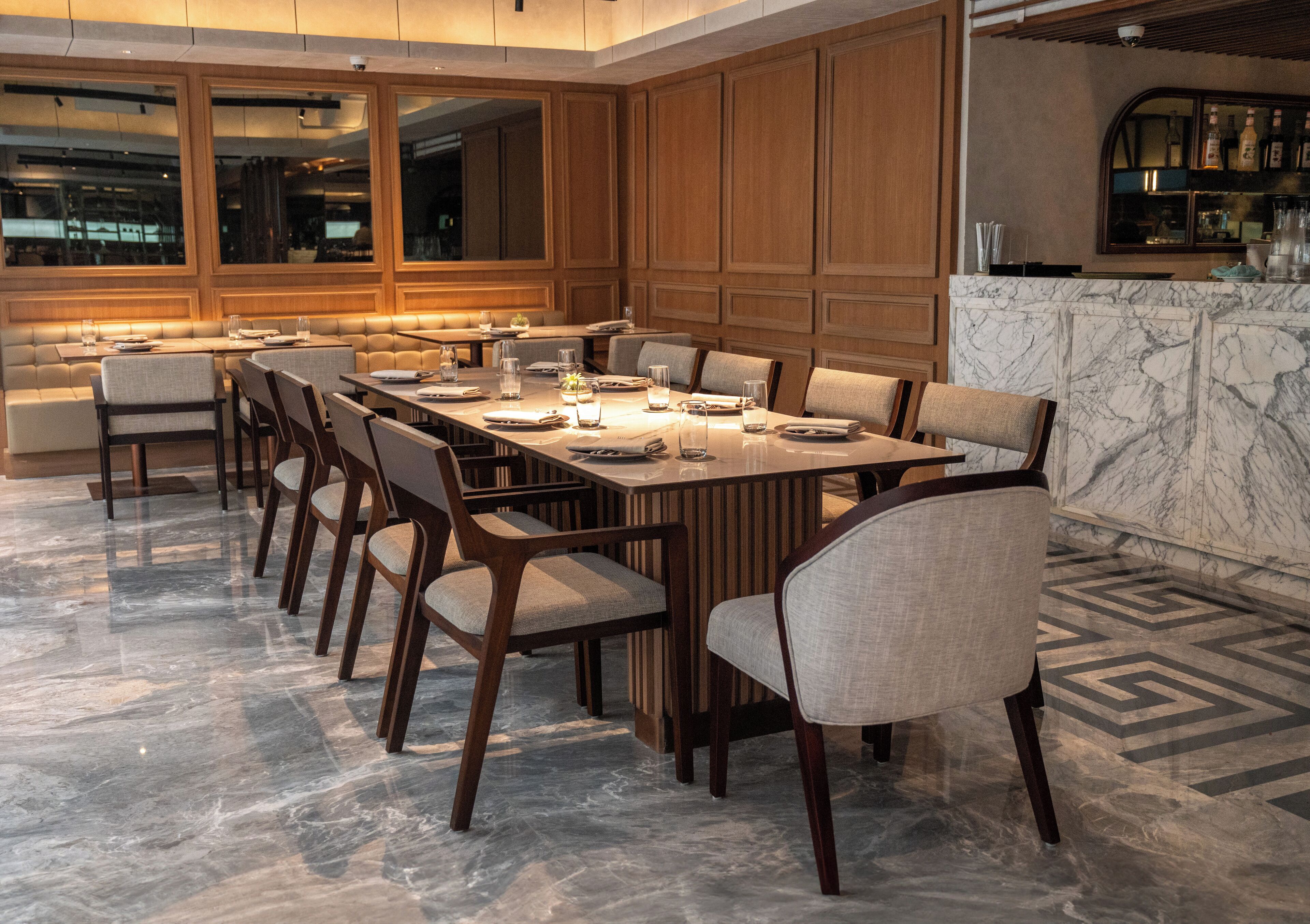 A modern dining area with a long table set for multiple people, wooden paneling on the walls, and a marble floor.