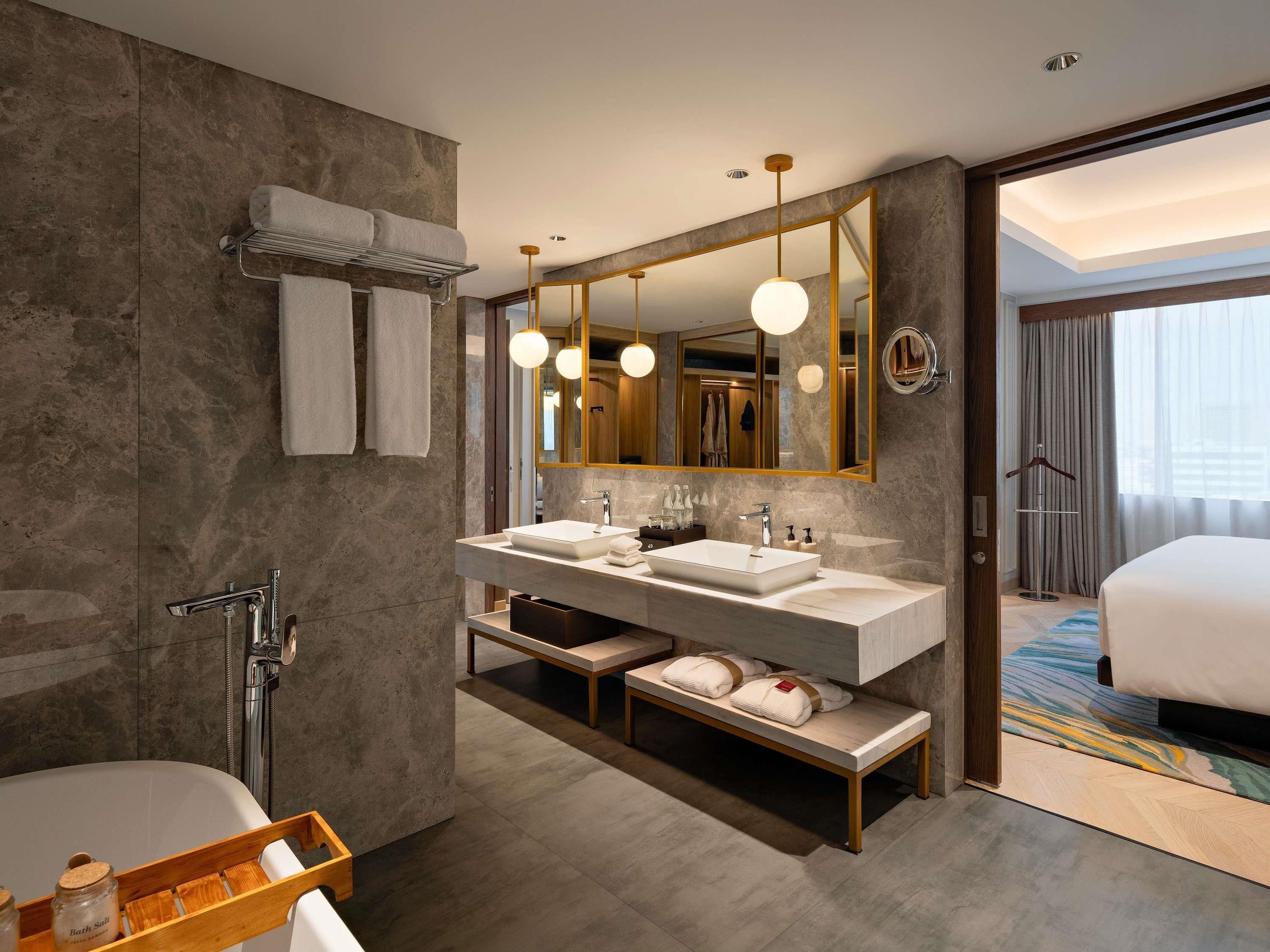 A modern bathroom with a large mirror, dual sinks, a bathtub, and a bench.