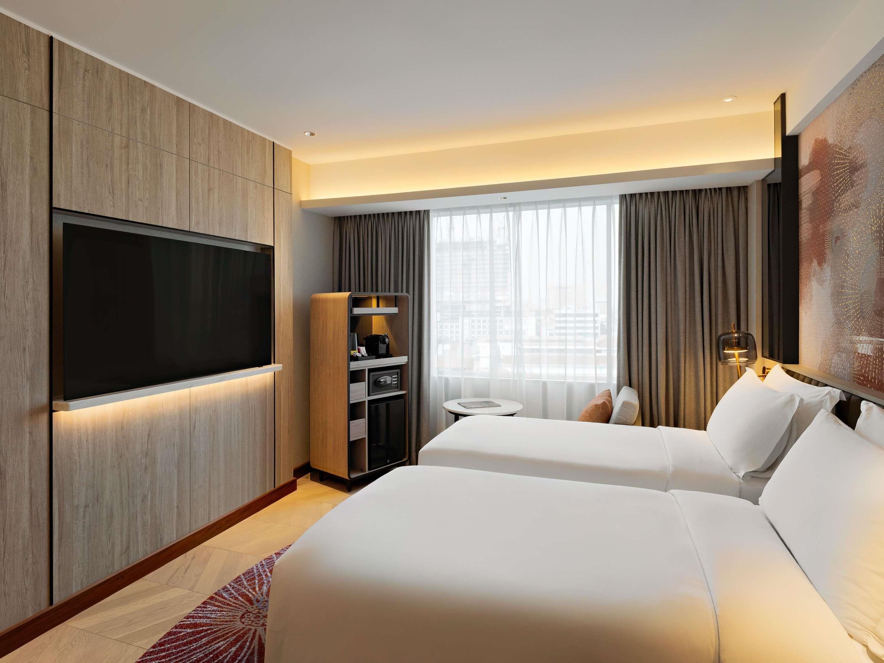A hotel room with a large flat-screen TV, a minibar, a small round table, and a bed with white linens.