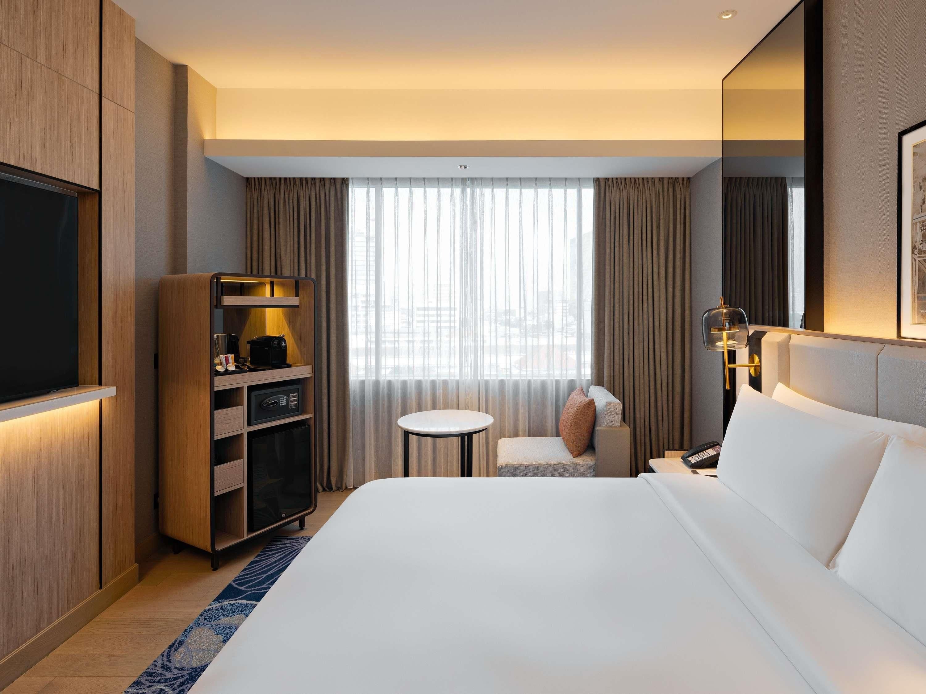 A hotel room with a large bed, a TV, a minibar, a seating area with a small table, and a large window with curtains.