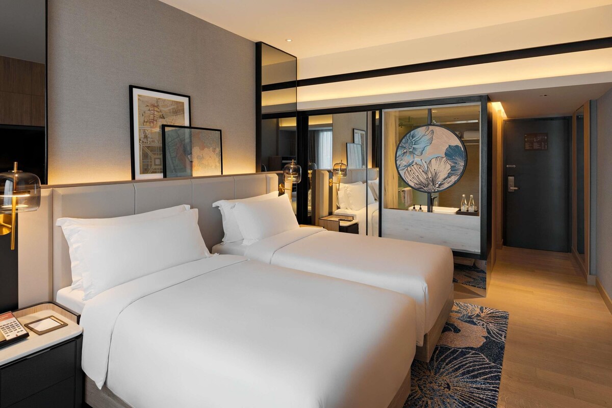 Habitación de hotel moderna con dos camas, un espejo grande, una mesita de noche con lámpara y vista al pasillo.