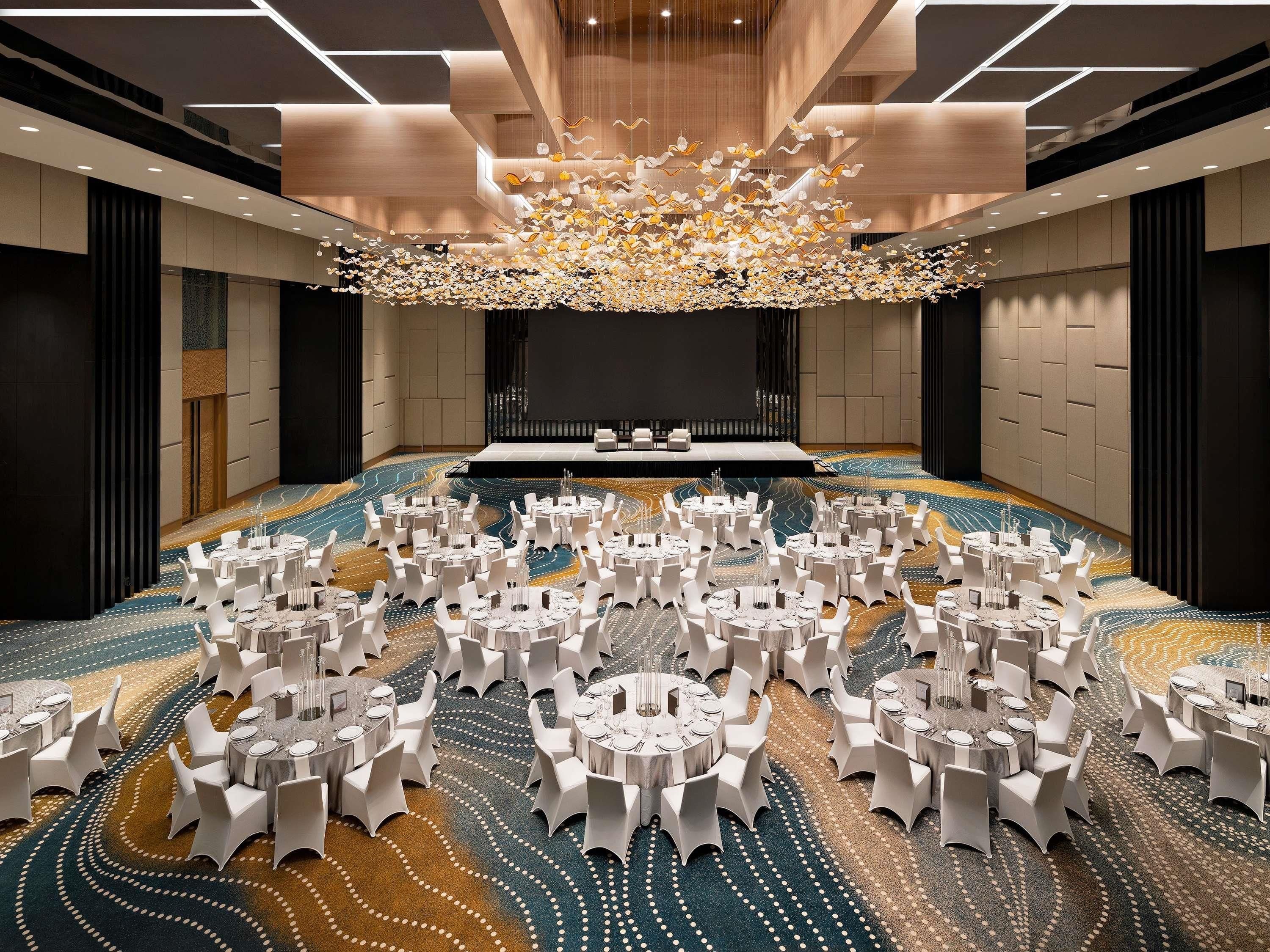 Een ruime congreszaal met ronde tafels en witte stoelen, een podium en een kamerbreed tapijt met een patroon.