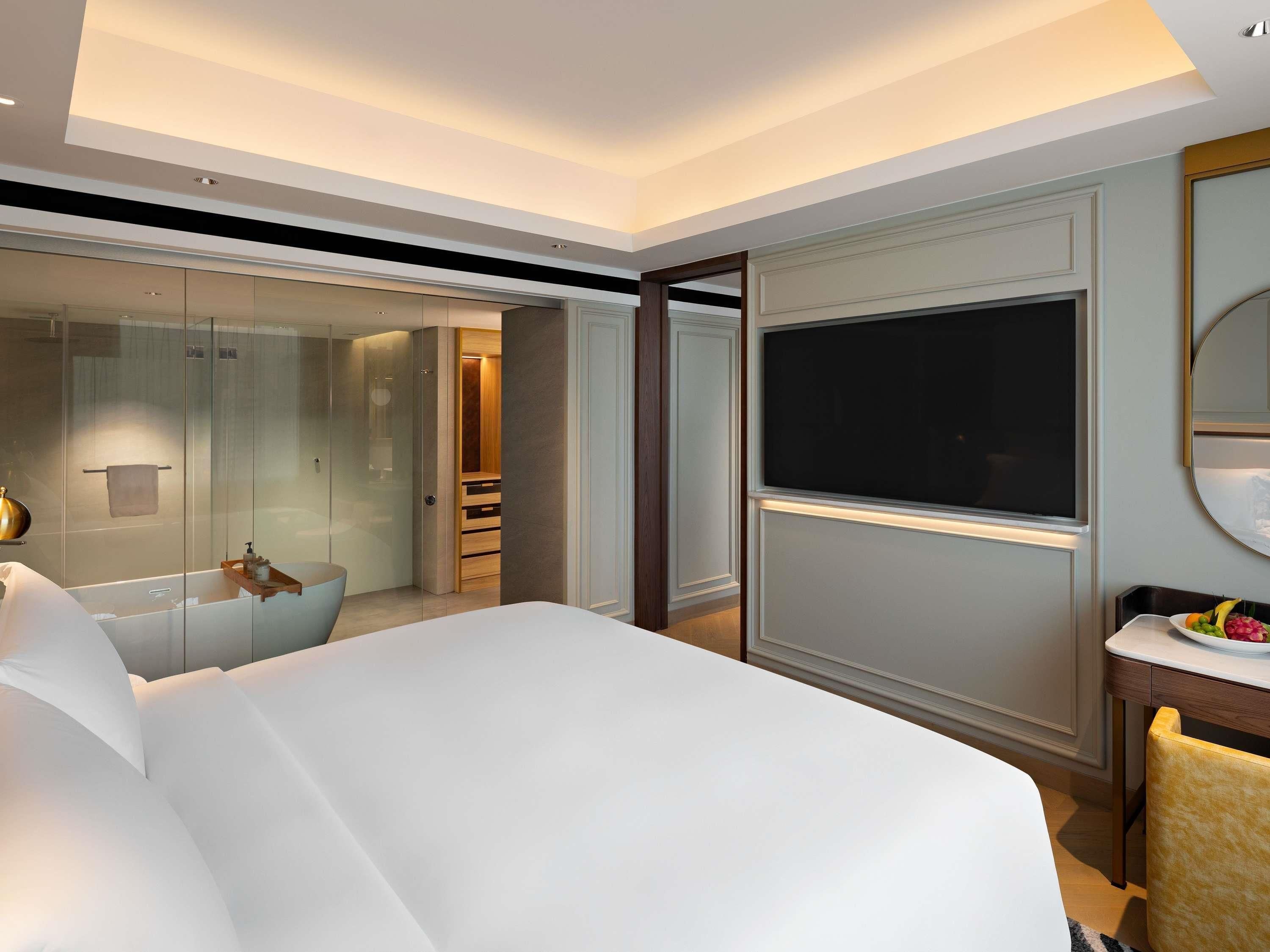 Ein modernes Hotelzimmer mit einem großen Bett, einer gläsernen Duschkabine, einem Flachbildfernseher und einem kleinen Esstisch mit Obst.