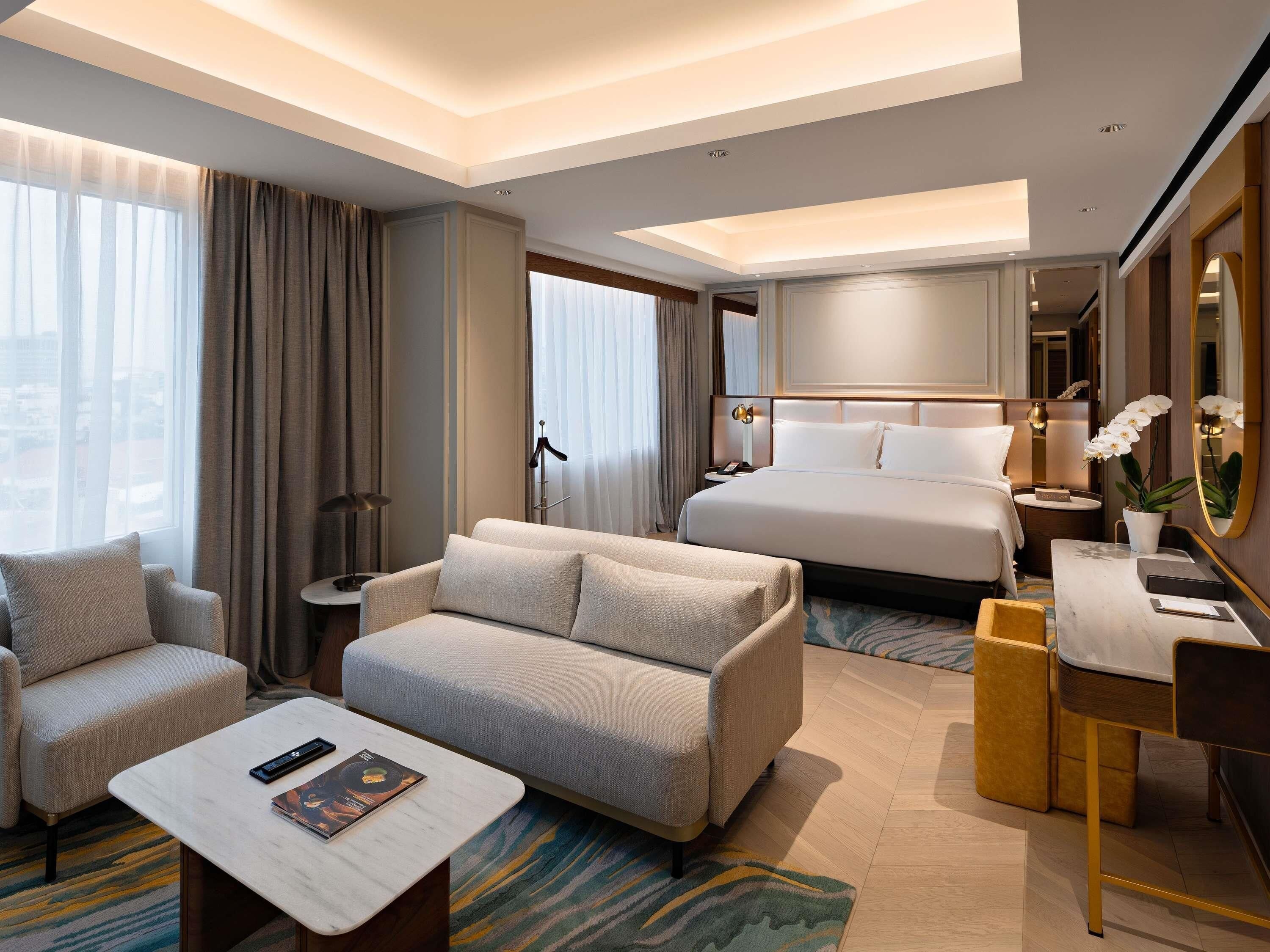 Une chambre d’hôtel comprenant un lit, un canapé, un bureau, une chaise, une table basse et une grande fenêtre avec des rideaux.