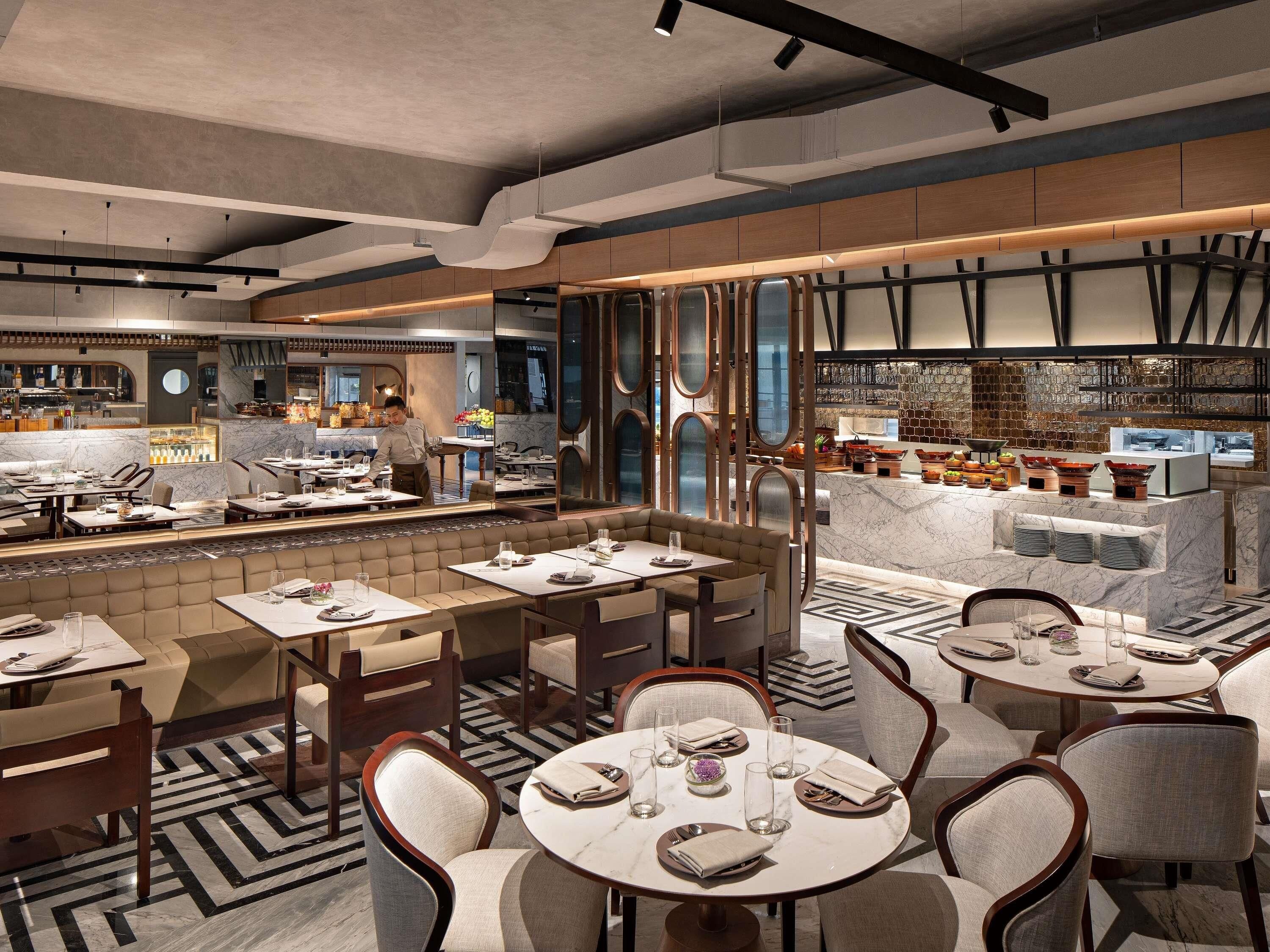 Um restaurante moderno com vários espaços de refeição, um bar central e um chef preparando alimentos.