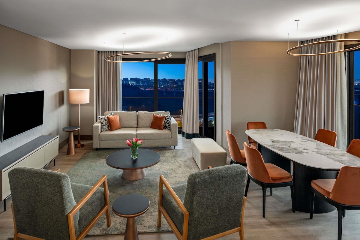 Ein modernes Hotelzimmer mit Sofa, Couchtisch, Esstisch und Stühlen. Es gibt einen Fernseher, eine Lampe und Blick auf die beleuchtete Stadt.