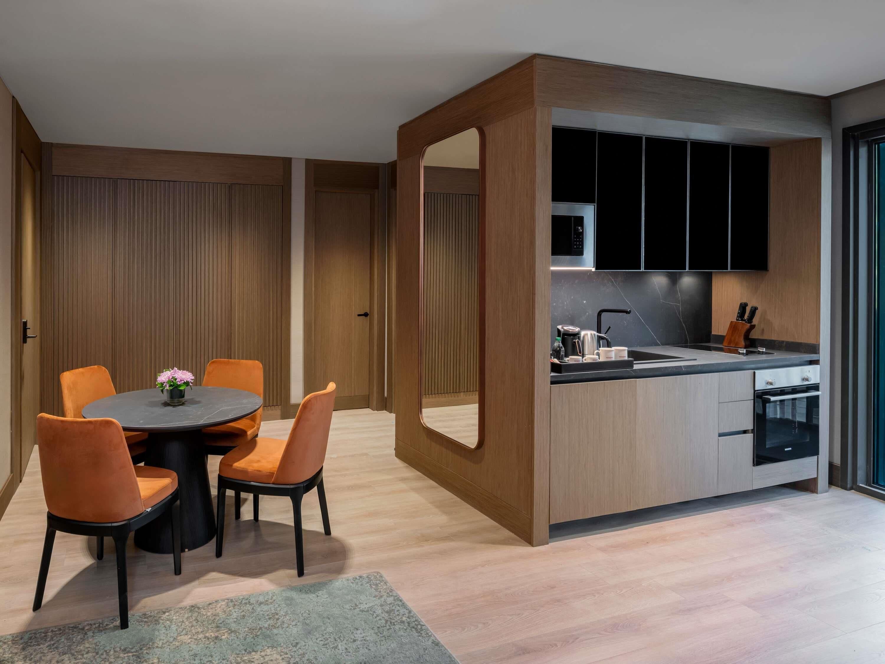 A modern kitchen with a central island, wooden cabinetry, and a dining area with orange chairs and a round table.
