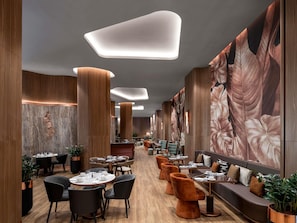 Een modern restaurant met houten lambrisering, grote hanglampen en een muurschildering van bladeren.