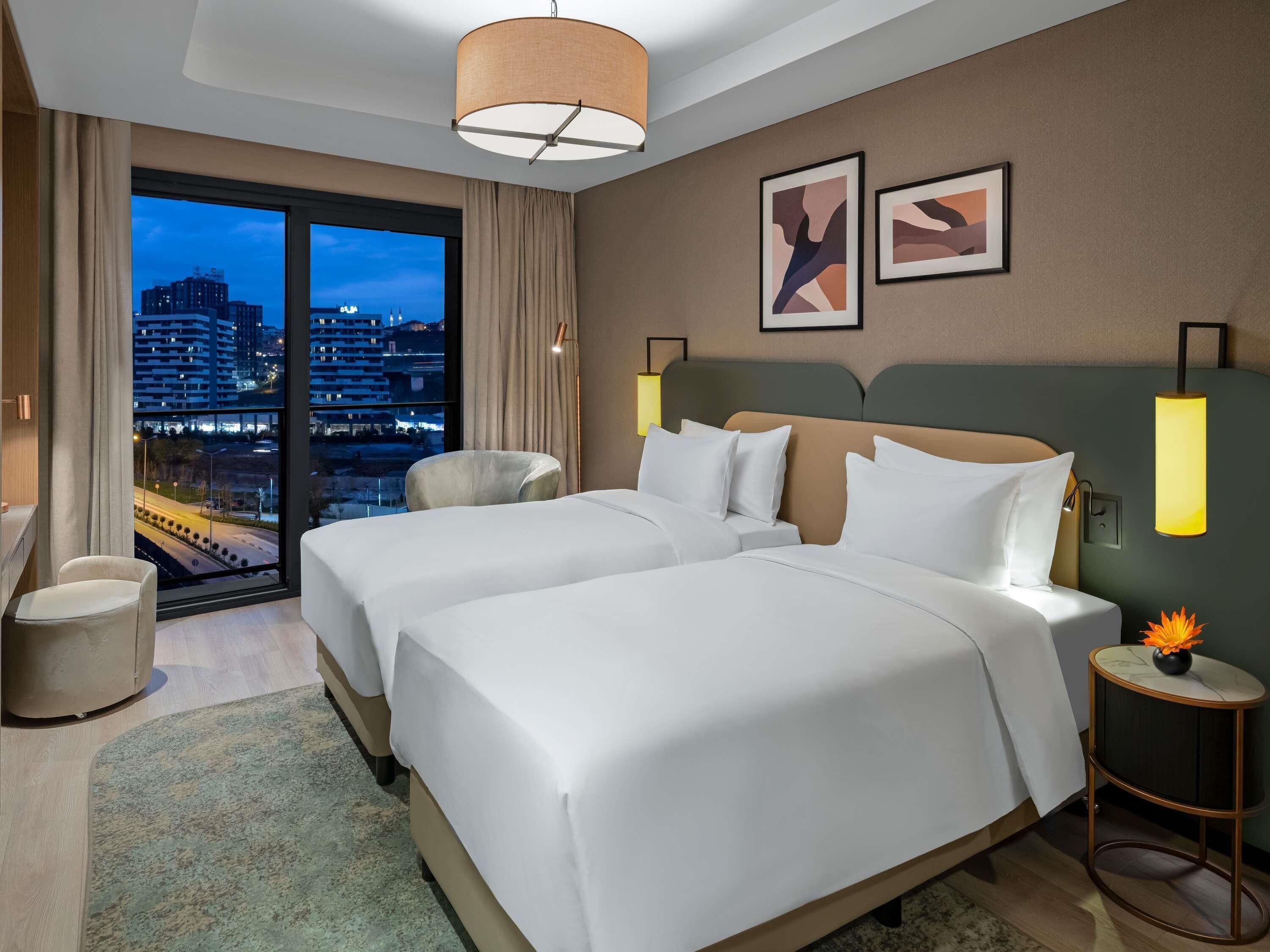 A hotel room with two beds, a view of a cityscape, a large window, and a nightstand with a lamp.