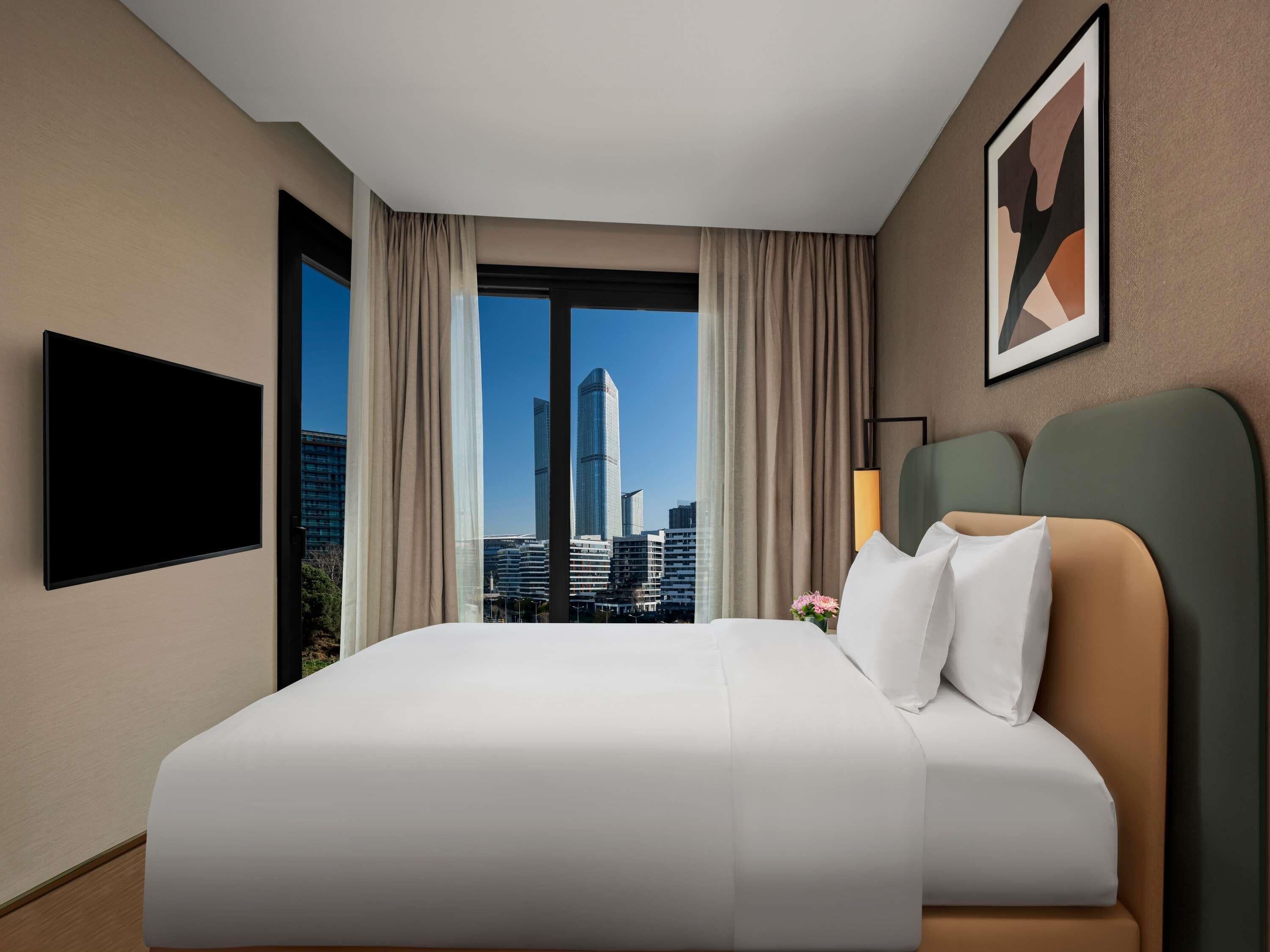 A hotel room with a large bed, a flat-screen TV, a city view, and a framed artwork on the wall.