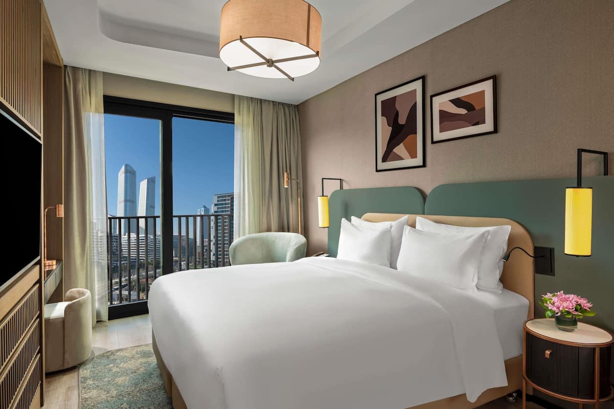 Hotelkamer met een groot bed, uitzicht op de stad, een balkon en een klein nachtkastje met een bloevas.