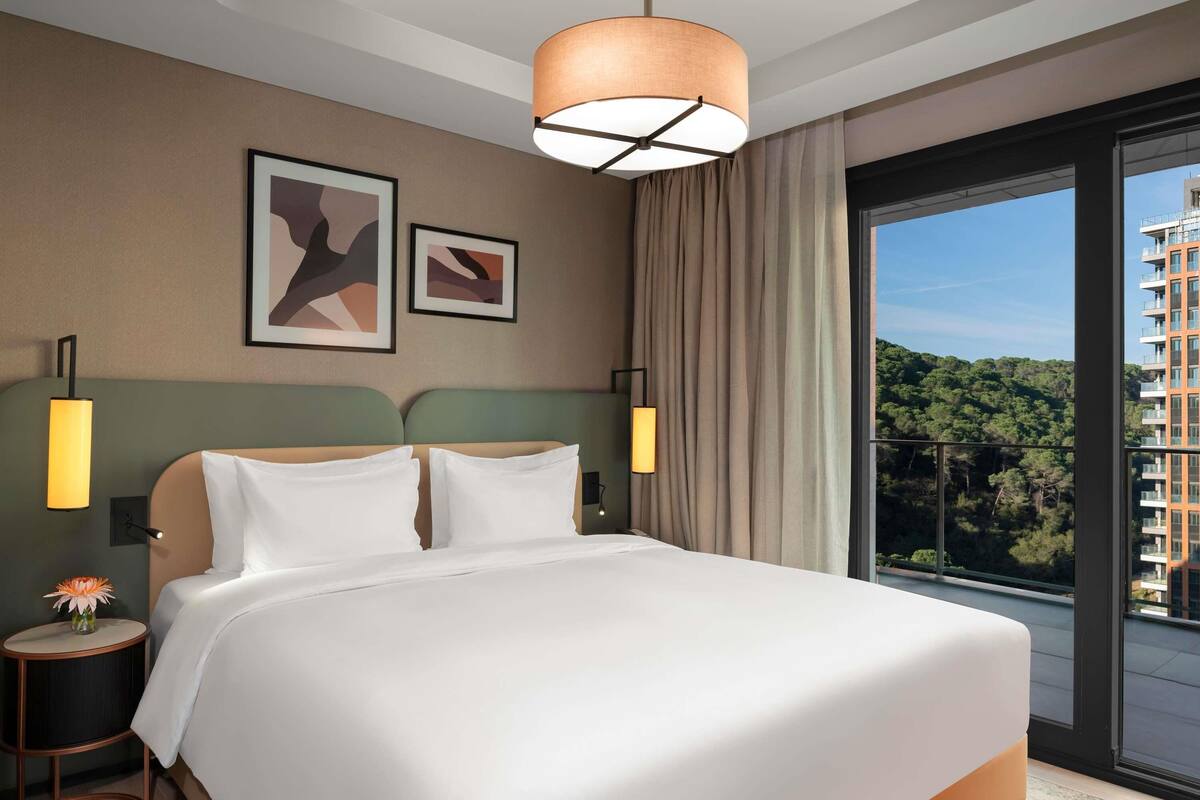 Hotelkamer met een groot bed, twee wandlampen en uitzicht op een gebouw en bomen door een groot raam.