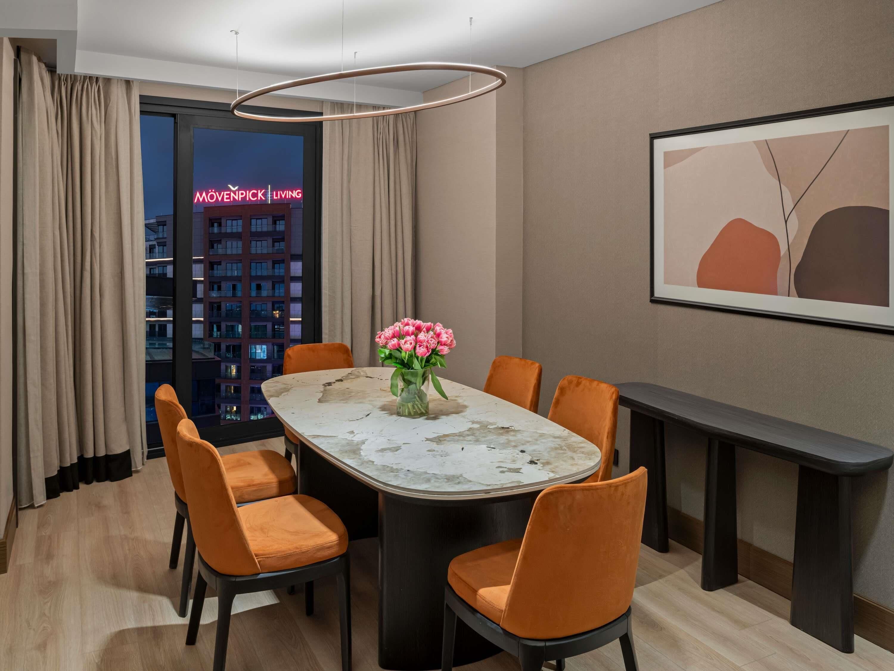 A dining area with a marble table, orange chairs, a vase of pink flowers, a modern pendant light, and a framed abstract painting on the wall.