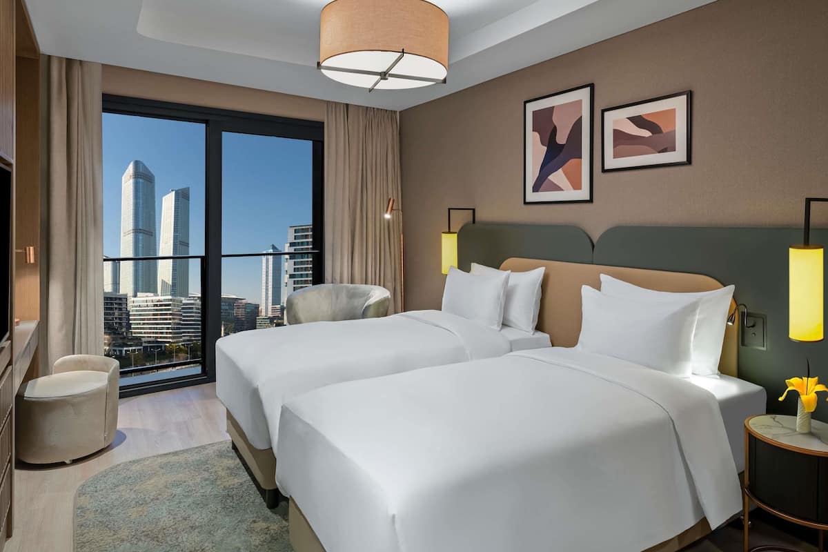 Een hotelkamer met twee bedden, uitzicht op hoge gebouwen, een stoel en een lamp.