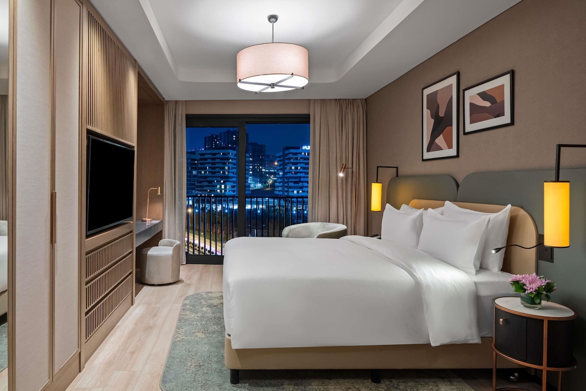 Ein modernes Hotelzimmer mit einem großen Bett, einem Fernseher, einem Balkon mit Stadtblick und einer Stadtansicht durch das Fenster.