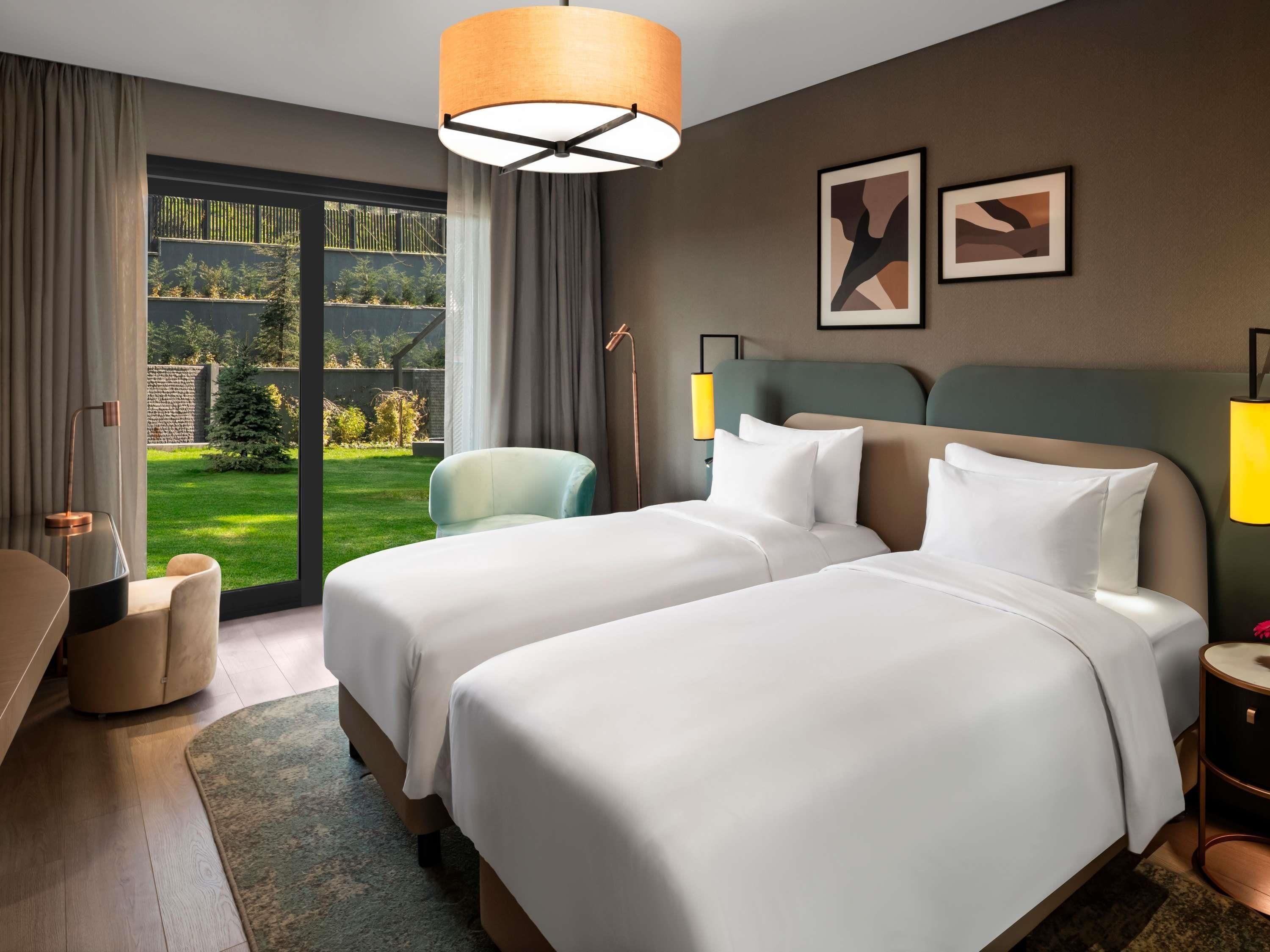 Habitación de hotel con dos camas, vista a un jardín y una lámpara moderna.