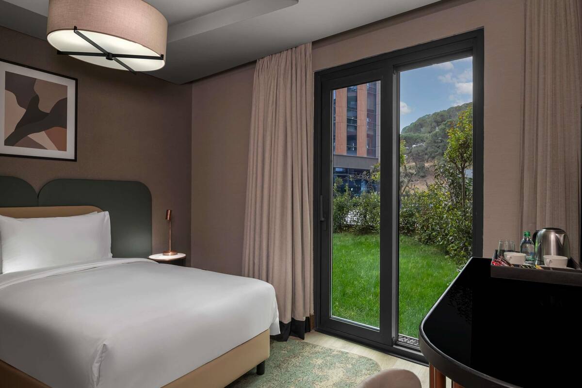 Hotelkamer met een bed, bureau, stoel, lamp, schilderij en uitzicht op groen en gebouwen.