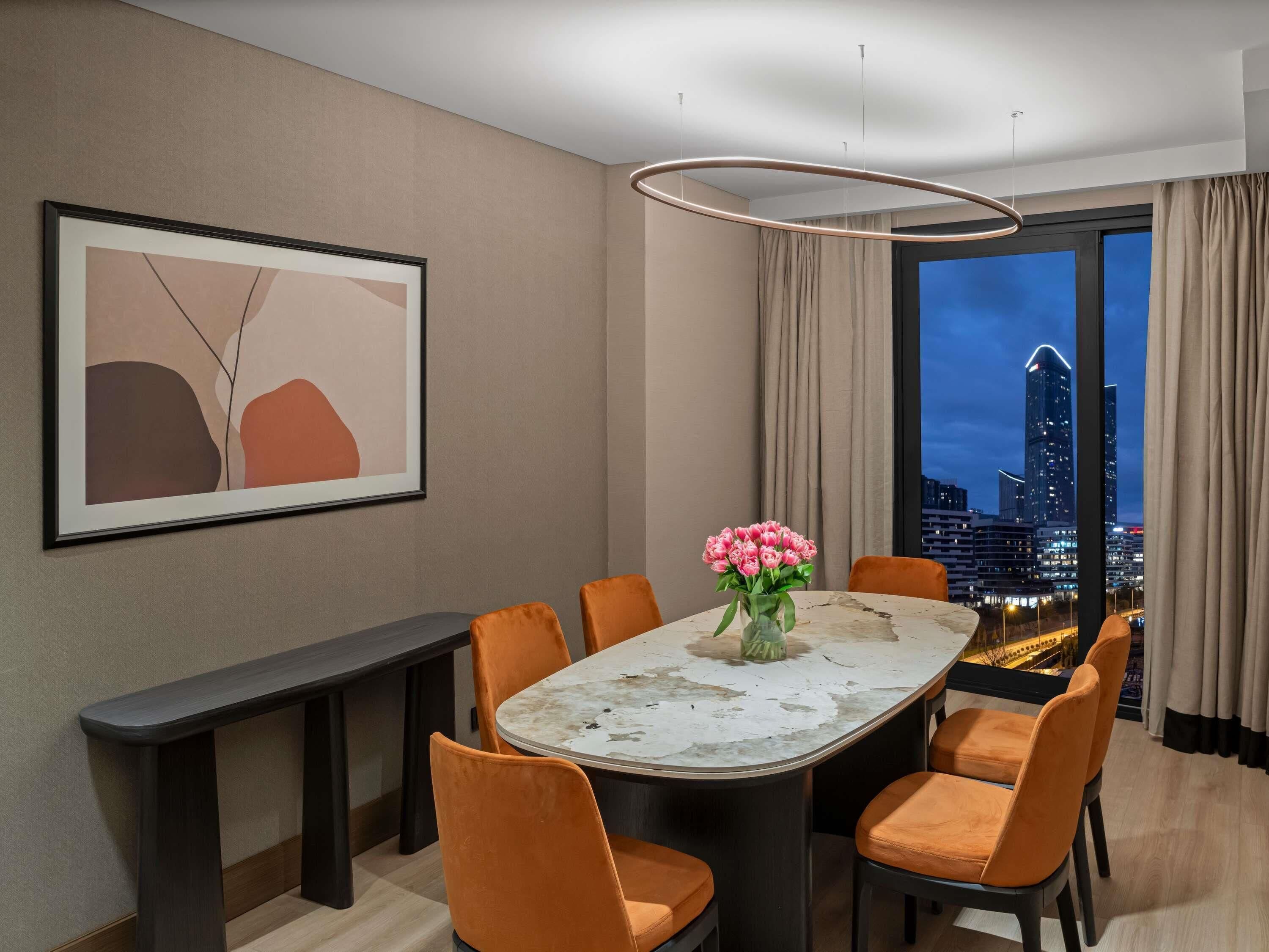 A modern dining room with a round table, orange chairs, a vase of pink flowers, a framed abstract painting, and a cityscape view through large windows.