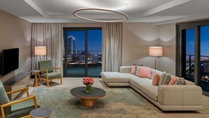 A modern living room with a sectional sofa, a flat-screen TV, a coffee table with flowers, and a city view through large windows. - Mövenpick Living Istanbul Saklivadi (Istanbul)