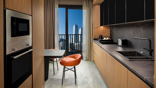 A modern kitchen with a built-in oven, microwave, and sink. There is a dining area with a table and chairs, and a view of a cityscape through a large window. - Mövenpick Living Istanbul Saklivadi (Istanbul)