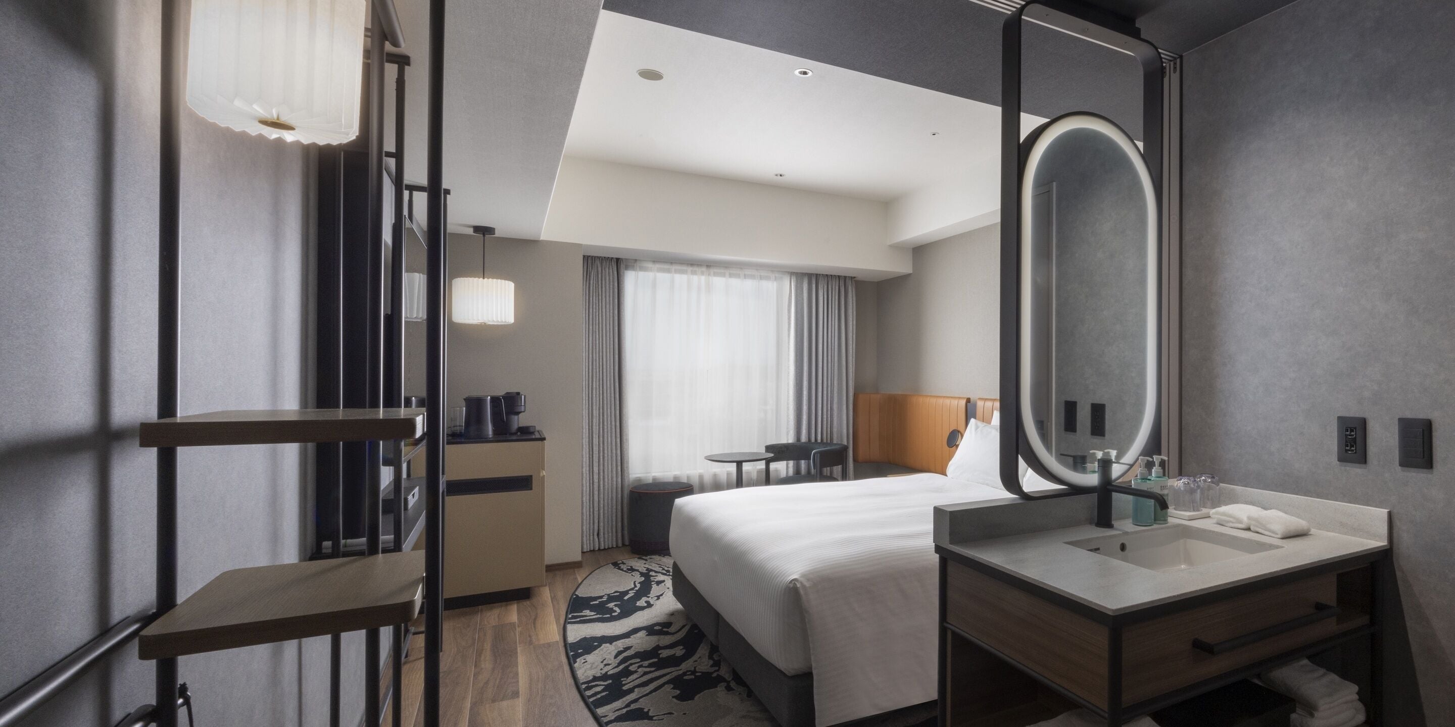 Habitación de hotel con cama, escritorio, silla, espejo redondo, lavamanos y ventana con cortinas.