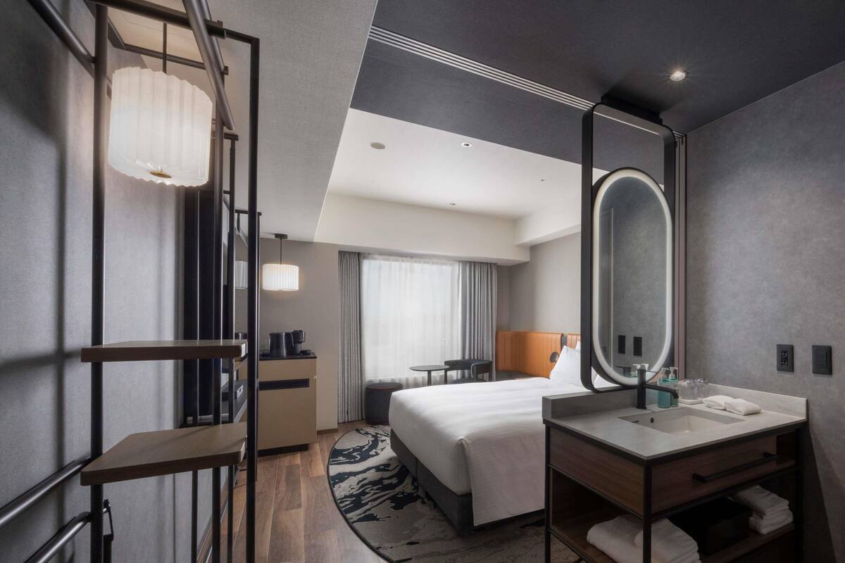 A modern hotel room with a bed, a desk, a chair, a bathroom with a mirror, and a large window.