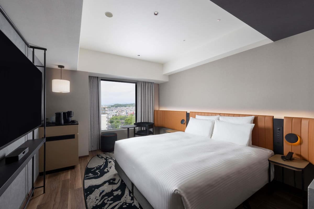 A hotel room with a large bed, a TV, a desk with a coffee maker, a small table, and a view of the city.