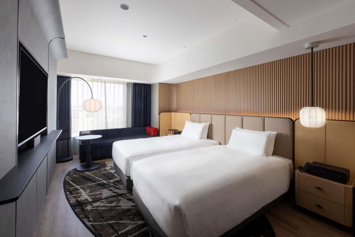 A modern hotel room with two beds, a flat-screen TV, a small round table, a sofa, and a patterned rug.