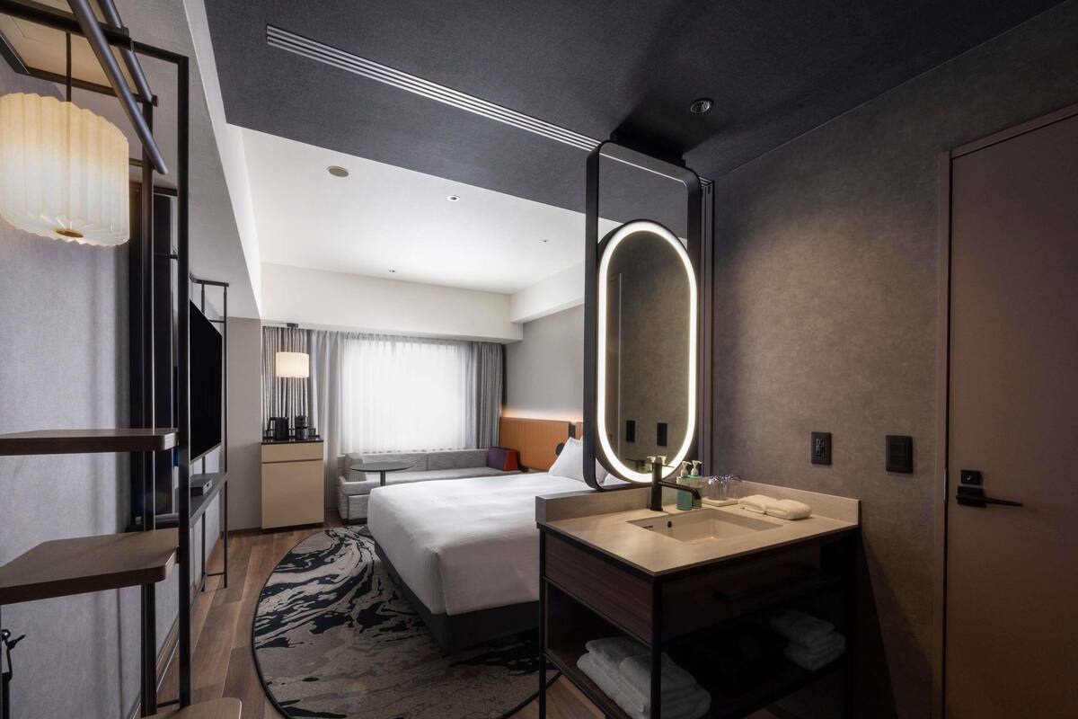 A modern hotel room with a bed, bedside table, mirror, and a small seating area.