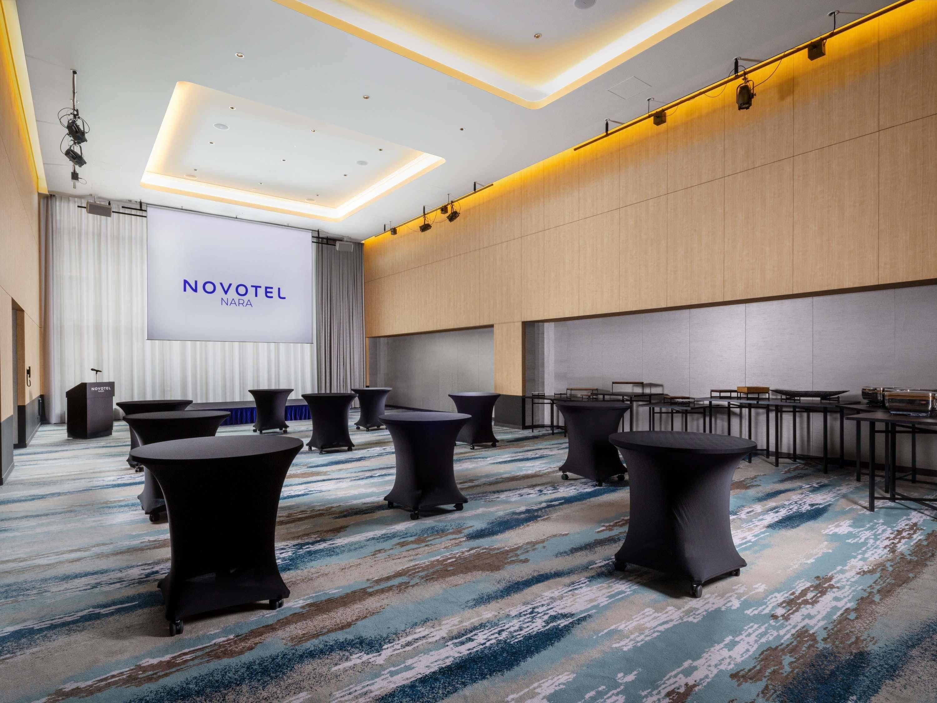 Une salle de conférence moderne dans un hôtel, dotée d’un grand écran affichant le texte « NOVOTEL NARA », de tables rondes et d’un pupitre.